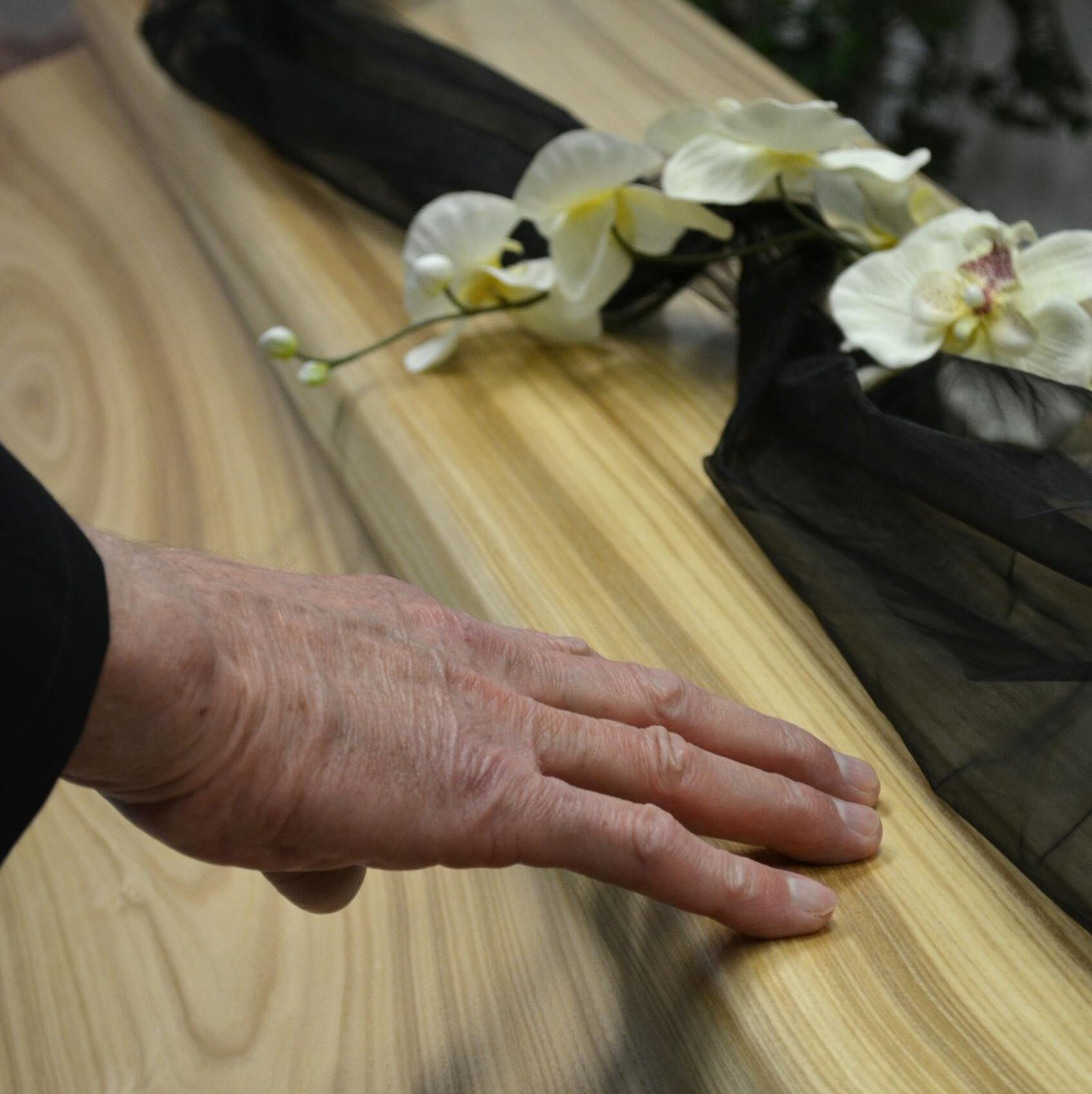 Close-up of a hand touching a wooden coffin adorned with flowers and black fabric, conveying a sense of mourning.